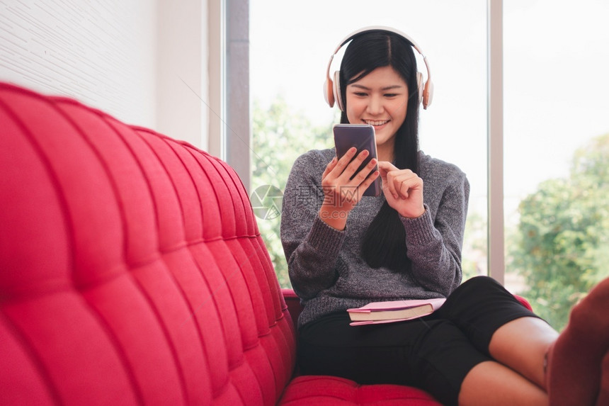 清晨坐在沙发上的亚洲可爱女人坐在沙发上用手机送信息同时听音乐在客厅大腿上放一本书笑声学习读白种人图片