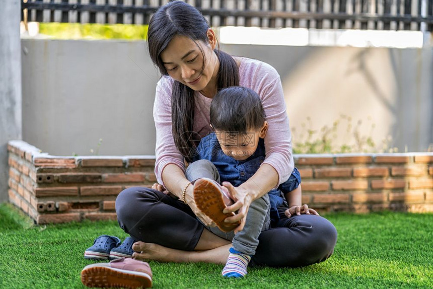 妈快乐的亚洲单身母亲在现代自学或家庭校和单身母亲概念选择重点的现代住房前草坪上为儿子穿亚洲单身母亲鞋活的