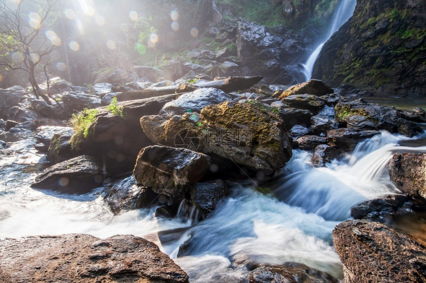 令人惊异的古老瀑布在夏天的景象明亮日光照耀在瀑布上温柔的水从溪流悬崖落下岩石和头中的红苔地衣抽象的bokoh背景泰国Khlong图片