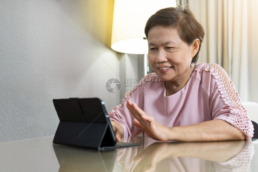 脸新冠国内的快乐笑亚洲年老妇女在家里隔离期间数字平板上进行视频呼叫身着粉红色衣服的老年妇女通过互联网技术与人们交谈时手挥同在流行图片