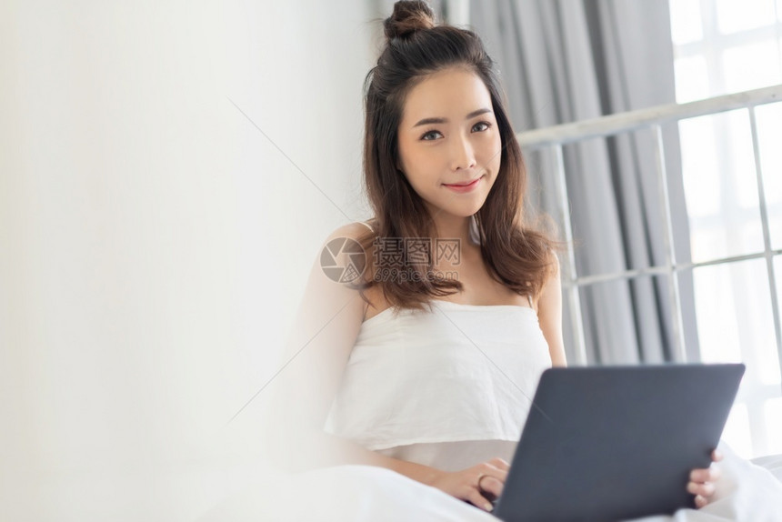 微笑早晨上坐在床笑着看镜头的漂亮女人肖像度假时在家白色卧室用笔记本电脑在白色卧室里使用亚洲美貌女孩阳幸福图片