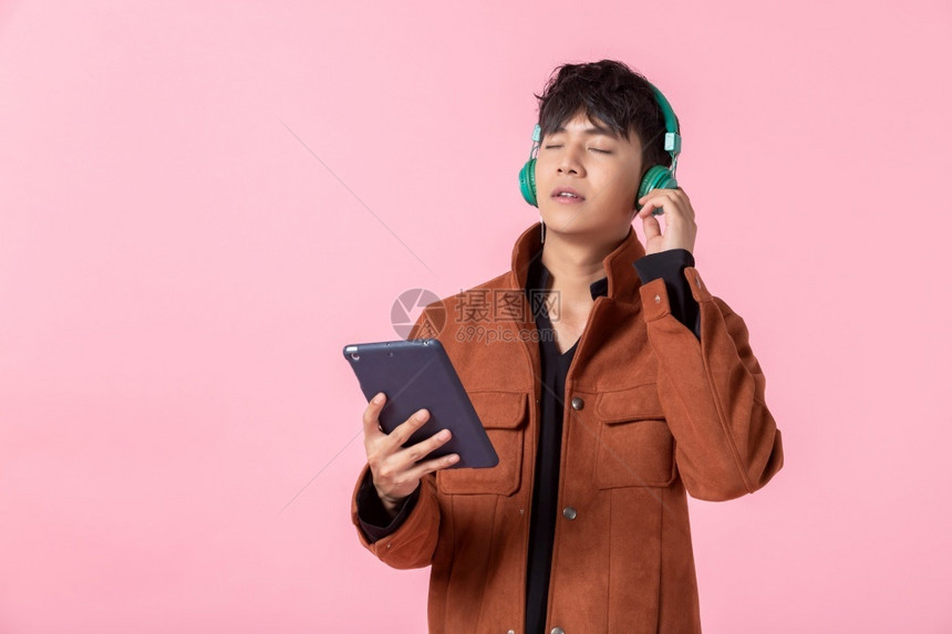 亚洲快乐一位在耳机上英俊的年轻男子在听音乐用平板电脑对侧眼双看镜头着爱中隔绝的相机拍着粉红色空白间工作室背景数字的积极男图片