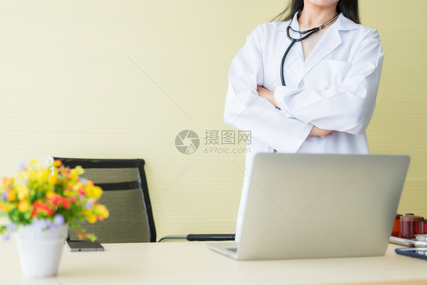 亚洲年轻女医生用听诊器和笔记本电脑将她的双臂围起在黄色医院背景中带有积极情绪的亚洲青年女医生紧关上实验室外套职业图片
