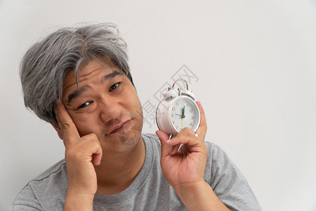 亚洲中年人举着一个白色闹钟脸闷不乐感觉很舒服他的问题是睡眠紊乱失是痛苦的念头无聊遭受成人坏的高清图片素材