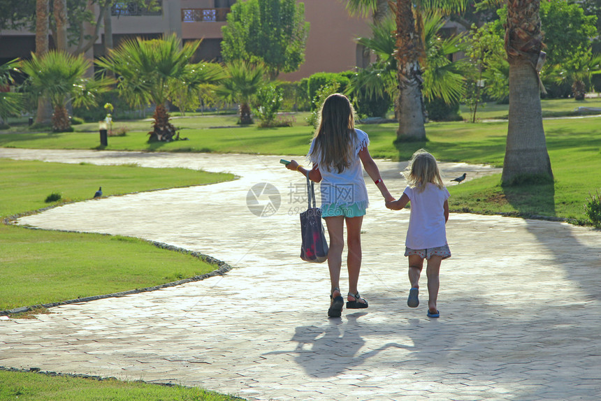 姐女士小旅游者在度假村的路上手握两个小姐妹在棕榈树中行走年轻女孩享受暑假子们放轻松概念孩子放轻松概念小旅游者在度假村的路上亲手拉图片