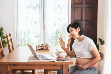 音你而乐毛笔字愉快沟通乐笑的亚裔年轻母亲在网上视频电话在她家怀孕时用笔记本电脑聊天和与家人握手时欢乐而美丽的孕妇享受肚子背景