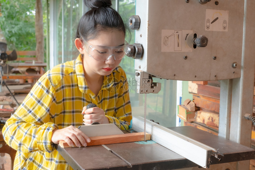 工程师站立妇女正在工作台上用木匠机带锯子的电动工具在作椅上手切割木材装修图片