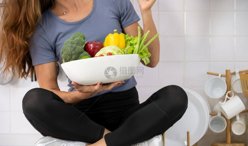 苹果有机的坐在厨房柜台上的年轻女士与运动服着新鲜蔬菜女运动员一起坐在厨房柜台手握白碗新鲜混合蔬菜健康食物和饮概念第6条图片