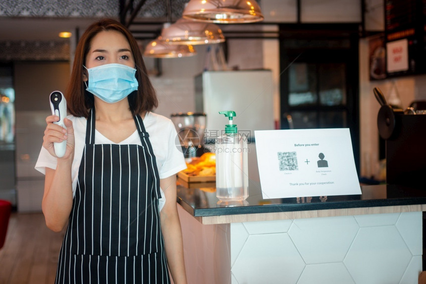 距离面具身戴罩和持有红外前额温度计的亚洲女服务员在进入餐馆咖啡店之前检查顾客身上的症状以检查体温在进入餐厅咖啡店之前检查客户的症图片
