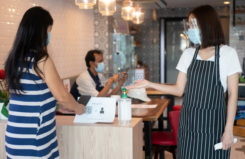 身戴面罩和持有红外温度计的亚洲女服务员建议顾客在进入餐馆咖啡店前使用手酒精消毒剂妇女还持红外温度计建议顾客在进入餐厅咖啡店前使用图片