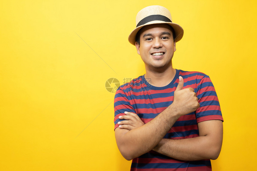 吸引人的模型穿着基本服装的快乐亚洲男子肖像穿着红色脱条T恤衫戴顶帽子微笑并展示拍相时伸出拇指显示成功与黄色背景隔绝图片