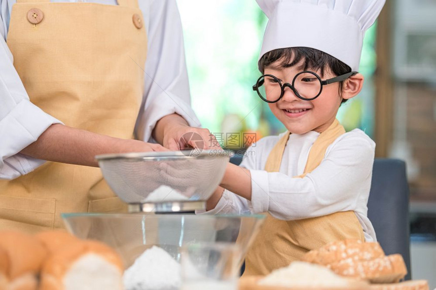 传统的家庭可爱亚洲小男孩和美丽的母亲在餐桌上的厨房里用筛选器把面粉擦成里有筛子细水沟在厨房里准备烘烤面包店和蛋糕微笑图片