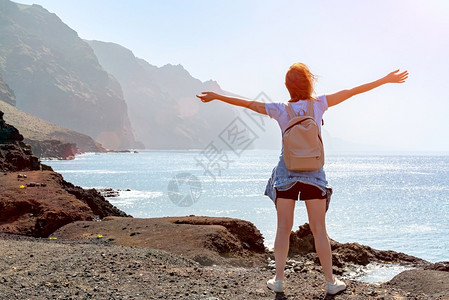 微笑一种背着包的年轻女子站在海洋岩石岸边靠近大海的女孩张开双臂在火山岩上迎接太阳光明媚的日子与亮蓝天在火山岩上迎接太阳光明媚的日女性高清图片素材