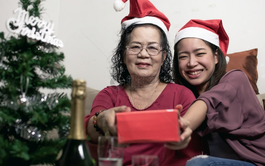两个60岁的老母亲和女儿戴着圣塔帽拥抱并持有礼物在家中带着模糊的树前景瓶装圣诞或新年庆典概念的干杯图片