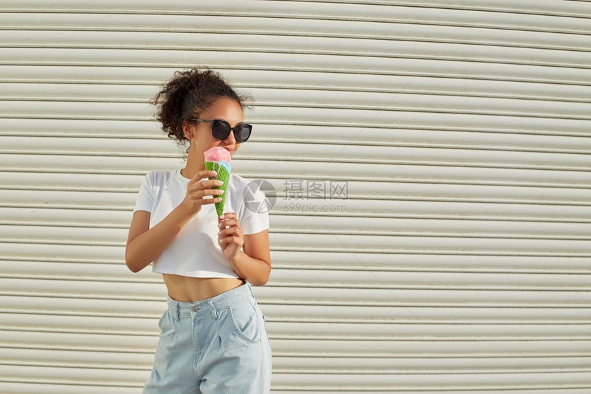 年轻的非裔美国人夏令穿着白色T恤衫和轻牛仔裤的年美籍非裔女孩在阳光日吃冰淇淋有选择地关注小重点领域图片