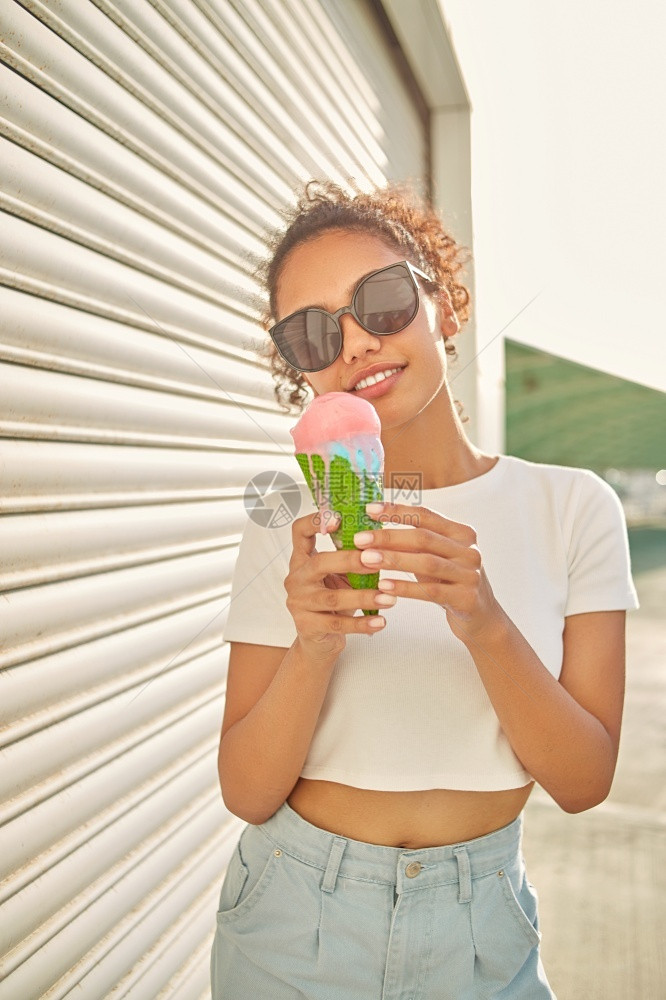 微笑穿着白色T恤衫和轻牛仔裤的年美籍非裔女孩在阳光日吃冰淇淋有选择地关注小重点领域甜的有趣图片
