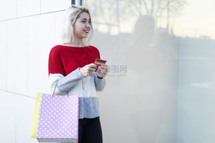 商业户外聪明的年轻消费女站在时装店橱窗露台前手持纸袋在阳光明日使用智能手机室外年轻消费女站在时装店窗露台前并使用智能手机校对Po图片