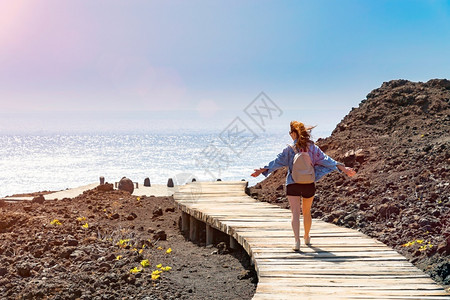 微笑散步欧洲背着包的年轻女子从岩石岸边的木平台上走到大海在阳光明媚的日子里在海边快乐的女人明亮蓝天特内里费岛的火山岩年轻女子背着背包高清图片素材