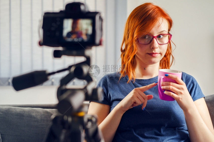 教程自由职业者年轻女caucasian女博客在家中或办公室制作视频博客的摄影机前展示茶杯或咖啡穿着T恤衫和眼镜看头前视互联网图片