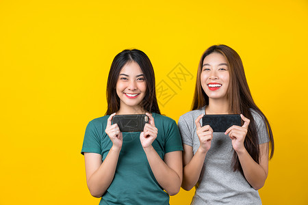 使用智能手机以孤立的黄色背景生活方式和业余爱好概念玩游戏的两幸福亚洲笑着年轻女子游戏家生活风格与休闲模型女失败者背景图片