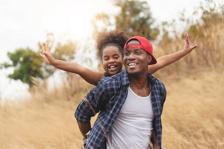 童年孩子快乐的黑人父亲微笑着玩得开心在秋天度假的非洲男人和裔美国女孩公园上给快乐的女儿搭便车享受干燥野地生活的家庭方式非洲人背景图片