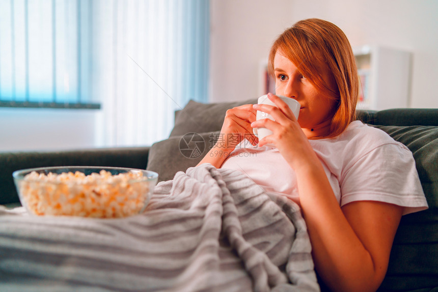 坐在家里沙发床上的年轻女士躺在一个爆米花碗里躺在一个爆米花碗上在她公寓独自享受电影或一系列喝茶咖啡的系列节目时看电视用毛毯子遮着图片