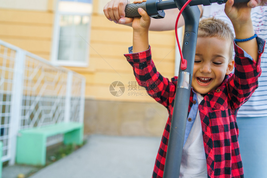 小快乐笑的男孩站在电踢推车上拿着风把手柄的摩托车由他母亲或姨妈其他女人驾驶常设保持户外图片