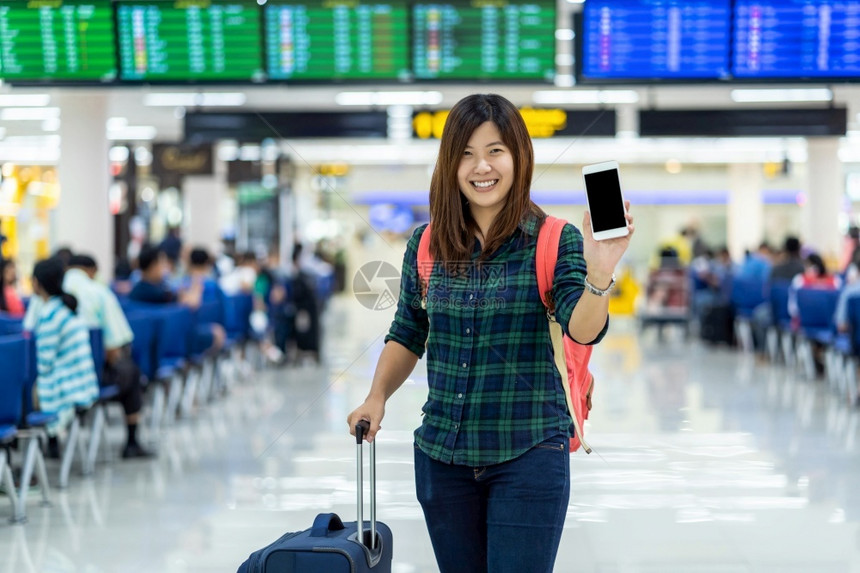 亚洲旅行者携带李在现代机场飞行信息屏幕的带有技术概念的旅行和运输上显示智能移动电话以登入飞行板的亚洲旅者和及运输工具挤移动的亚洲图片