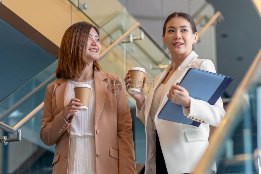 常设愉快女在现代办公室或合用场所的咖啡休息期间两名女亚洲企业家在咖啡休息期间散步和说话咖啡休息下班后放松和谈话商业人的伙伴关系概图片