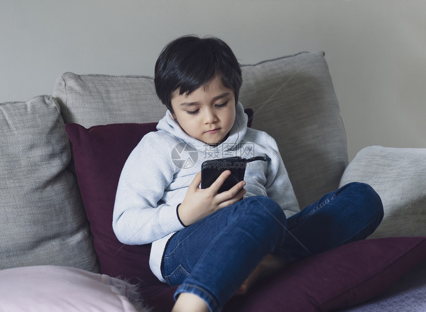 观看电话坐在沙发上看手机卡通片的男孩在校儿童放学时使用手机在互联网上学习教训的校儿童在家上学的庭就远程习在线教育概念疾病图片