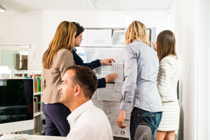 人们30多岁女在办公室工作的商界人士其中一些人看着公告板讨论贴在公告板上的设计另一个人在前面的正与几个计算机屏幕一起工作混合的c图片