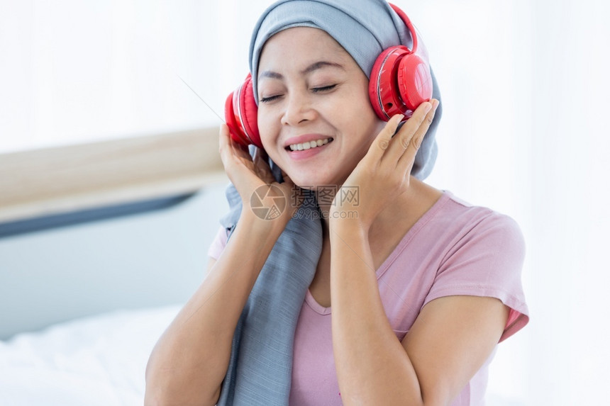 脸一位患有乳癌的亚裔女患者快乐她带着粉红色丝在耳机上戴头巾正在听音乐治疗后化就坐在床上房子里的卧室快乐亚洲人图片