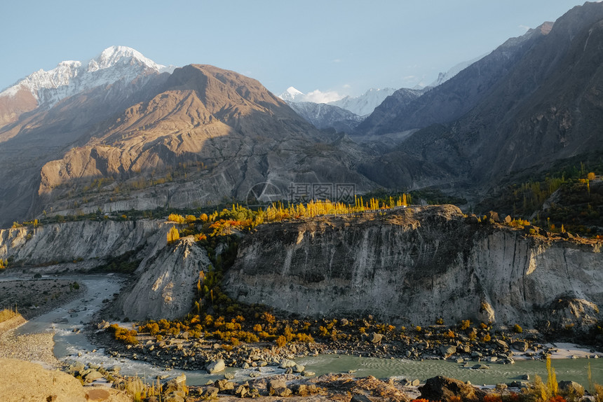 在巴基斯坦GilgitBaltistan的秋天卡拉科姆山脉HunzaNagar山谷的雪盖峰上照耀多彩树木和河流的自然阳光树叶平静图片