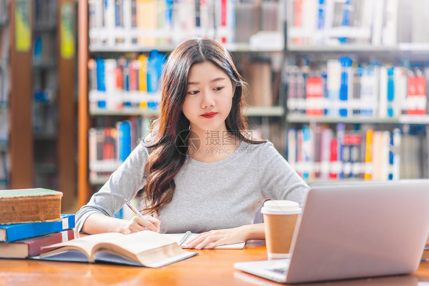 做功课和在大学图书馆或使用技术笔记本电脑的亚洲青年学生或具有各种书籍和文具架背景的各种群落返回学校从事家庭作业和使用技术笔记本电图片