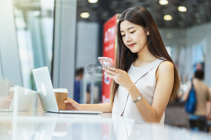 手机杯子亚洲年轻女持有一杯咖啡在现代店或合用空间使带有技术笔记本电脑的移动话技术智能移动和潮时生活方式企业家概念等亚洲青年女妇拥图片