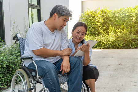 医院爱坐在轮椅上妻子坐在旁边并拿着平板电脑病人因车祸而坐轮椅妻子在旁边让他花园漫步时放松一下不要让他看到与肩并和鼓励的观念药片图片