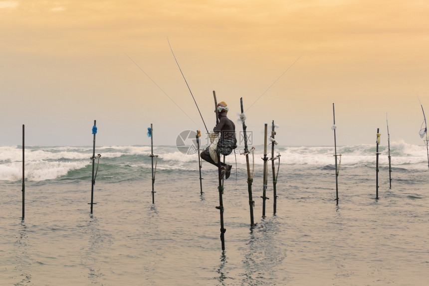 在日落晚夕坐杆子上手拿着一根木钓鱼棍的矮渔夫坐在他身后深处的海浪在幕后坠落天空碰撞橙图片