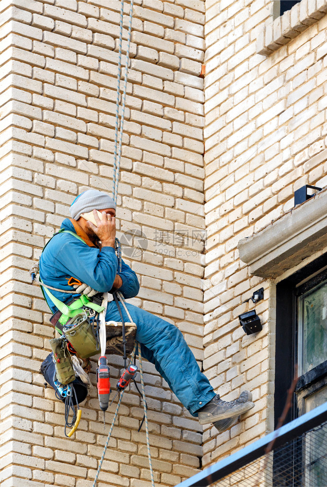 工业的窗户一种名工业登山者在旧建筑的砖墙上挂着安全绳用一系列各种工具捆绑在背后并收到电话指示一张将空间建造者攀爬吊在老砖楼墙上的图片