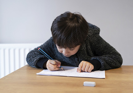 男生纯字素材学校男孩坐在桌边做功课快乐儿童拿着铅笔写作一个男孩在白皮书小学和家庭教育概念上写英语字的画像男孩坐在桌上做功课男孩在白皮书上写英背景