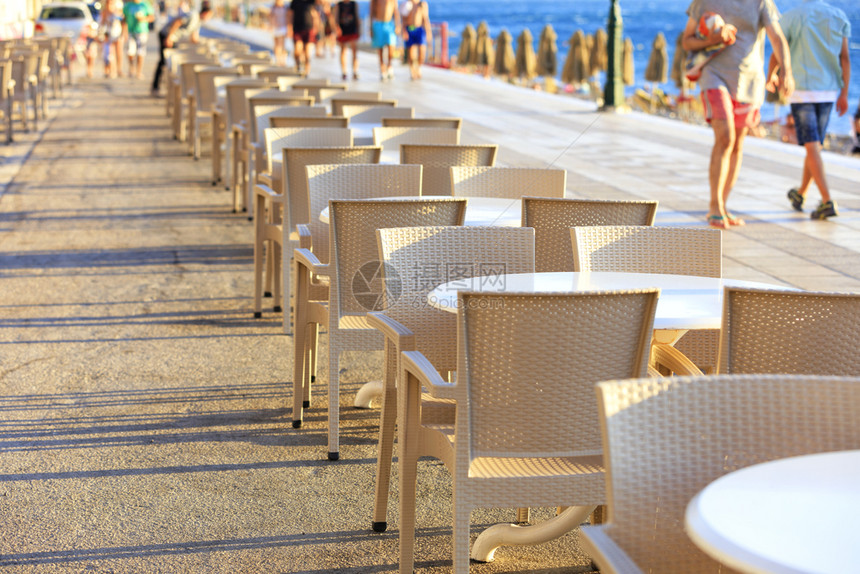 海滨滩咖啡厅的餐桌和椅子位于度假者行走的序道沿线海滩咖啡厅的桌和椅子位于游荡人群和蓝海中沿途散步的阶道一带蓝色的人们图片