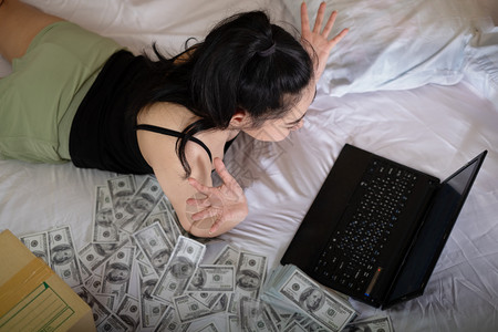 微笑快乐的淑女躺在床上卧室的笔记本电脑上工作里面有10美元的钞票很多来自家概念的作品以盈利为目的卖商品SellProfessio利润高清图片素材