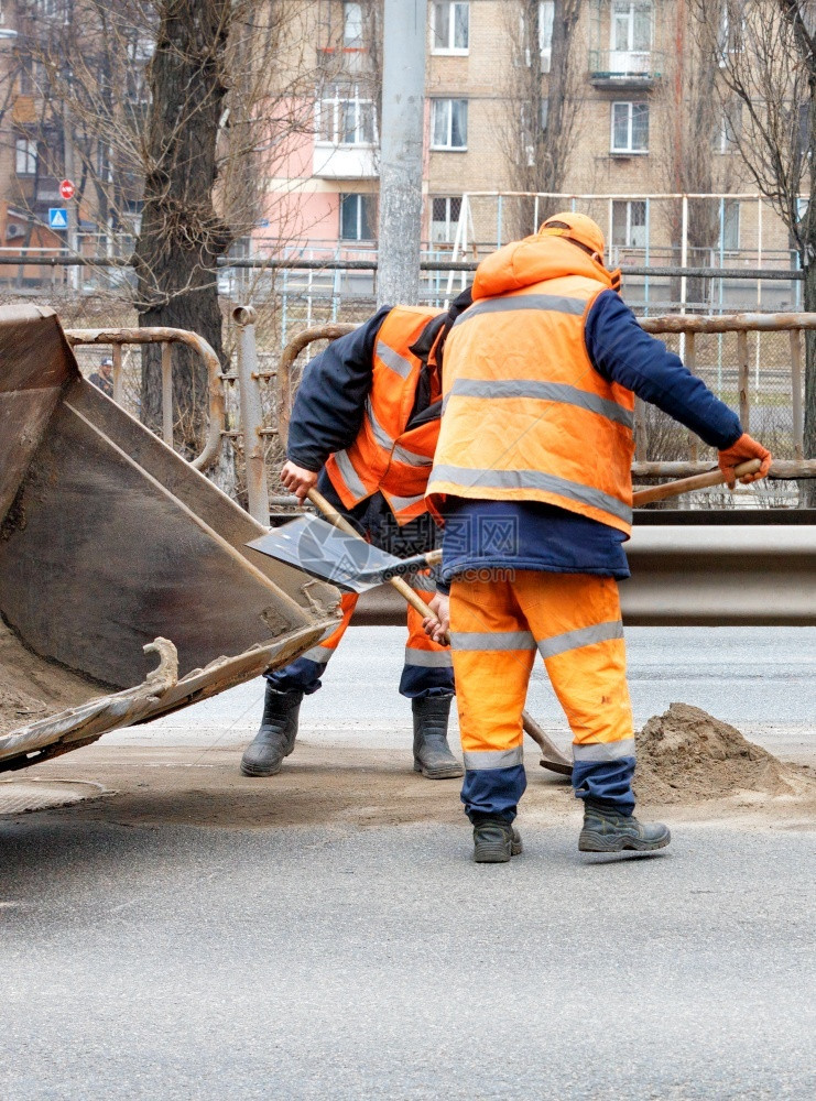 行业身穿明橙色反射制服的道路工人使用铲子清除各行道间的残块并将其装入一个平板桶垂直图像中道路工人将之间积聚的肮脏沙子铲到平板桶垂图片