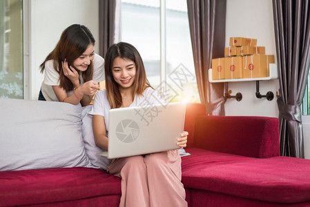 两名年轻的自由职业青年亚洲女企家在庭办公室私人工作手持膝上型计算机坐在沙发包装网上购物服务和向客户推销广告的网上邮包交付服务以及买高清图片素材