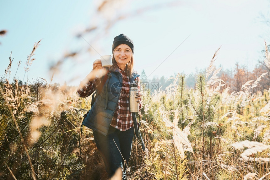 度假旅行期间在阳光明亮温暖的下享受咖啡的妇女站在高草地上的女人看着照相机拿咖啡杯和热水瓶子女人背着包徒步走在山中与自然相近的休闲图片