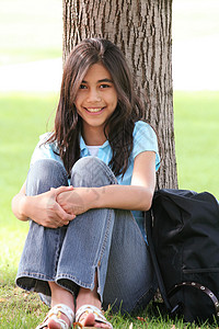 年轻女孩坐在树边坐着青少年公园草地学生混血中学背包微笑孩子背景图片