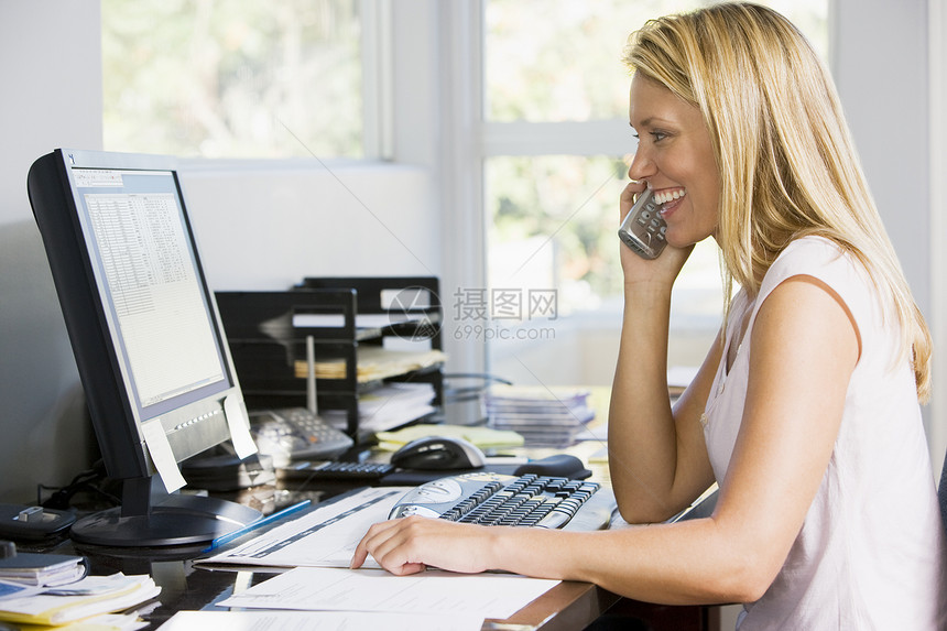 使用电话微笑的计算机在家庭办公室用电脑工作的妇女图片