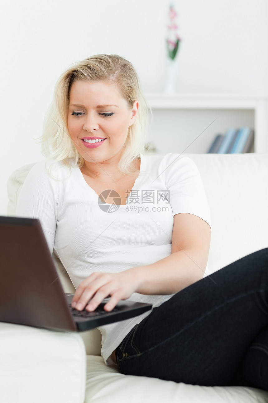 使用笔记本电脑坐在沙发上金发白色长椅休息室闲暇快乐金发女郎互联网微笑客厅图片