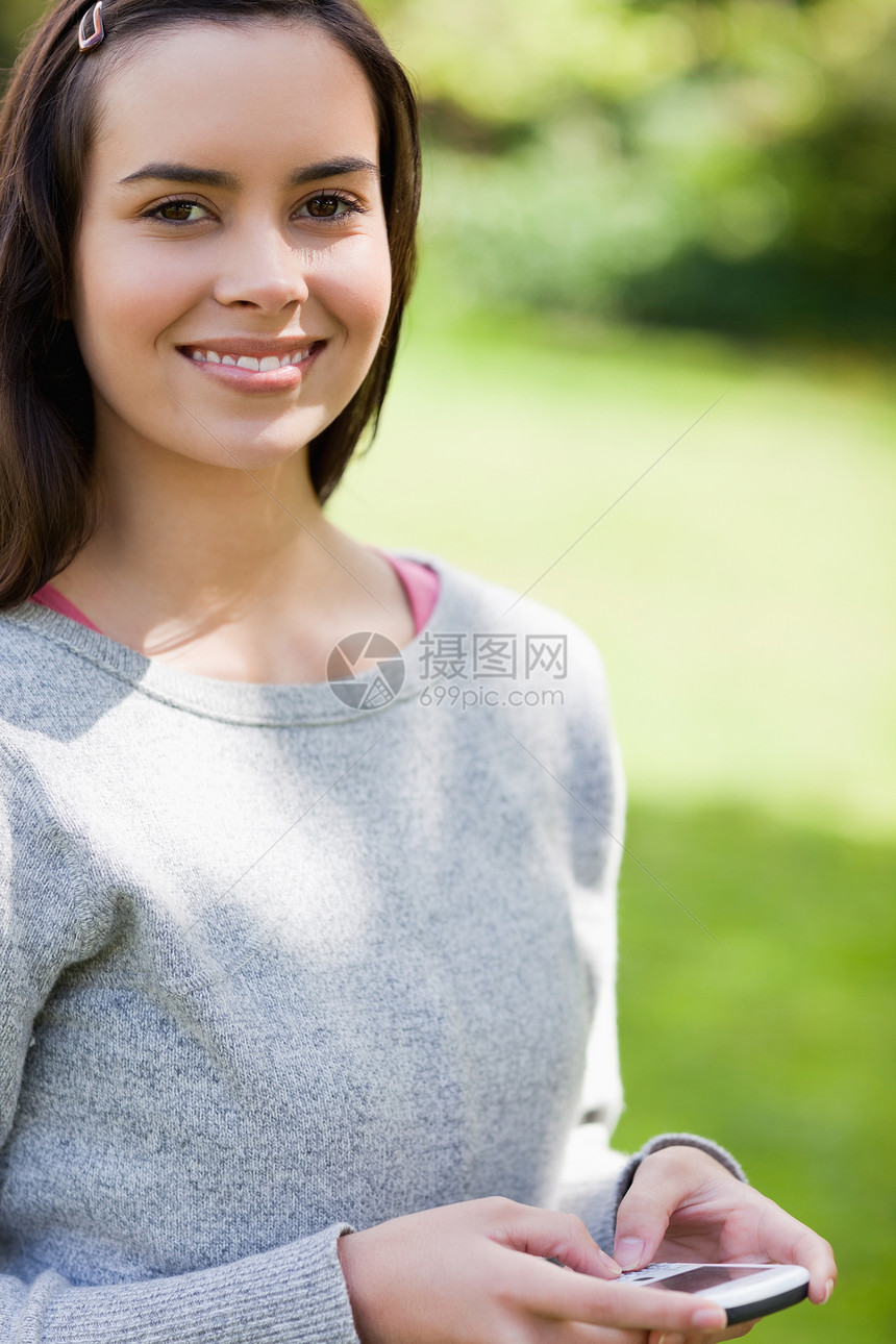微笑的年轻女子站在公园里时使用她的手机图片