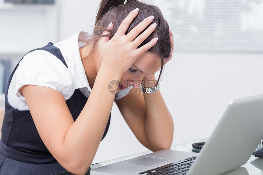 在书桌笔记本电脑前头握着头的沮丧女商务人士图片