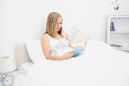 妇女在床上阅读书枕头女性床头柜家庭生活金发金发女郎闹钟卧室架子棉被背景图片